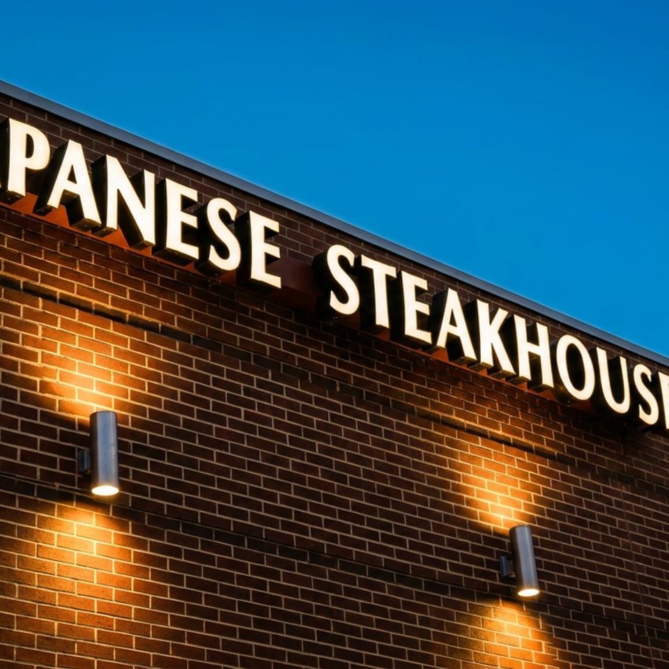 Illuminated "Japanese Steakhouse Sushi" sign on a brick building under a clear blue sky.