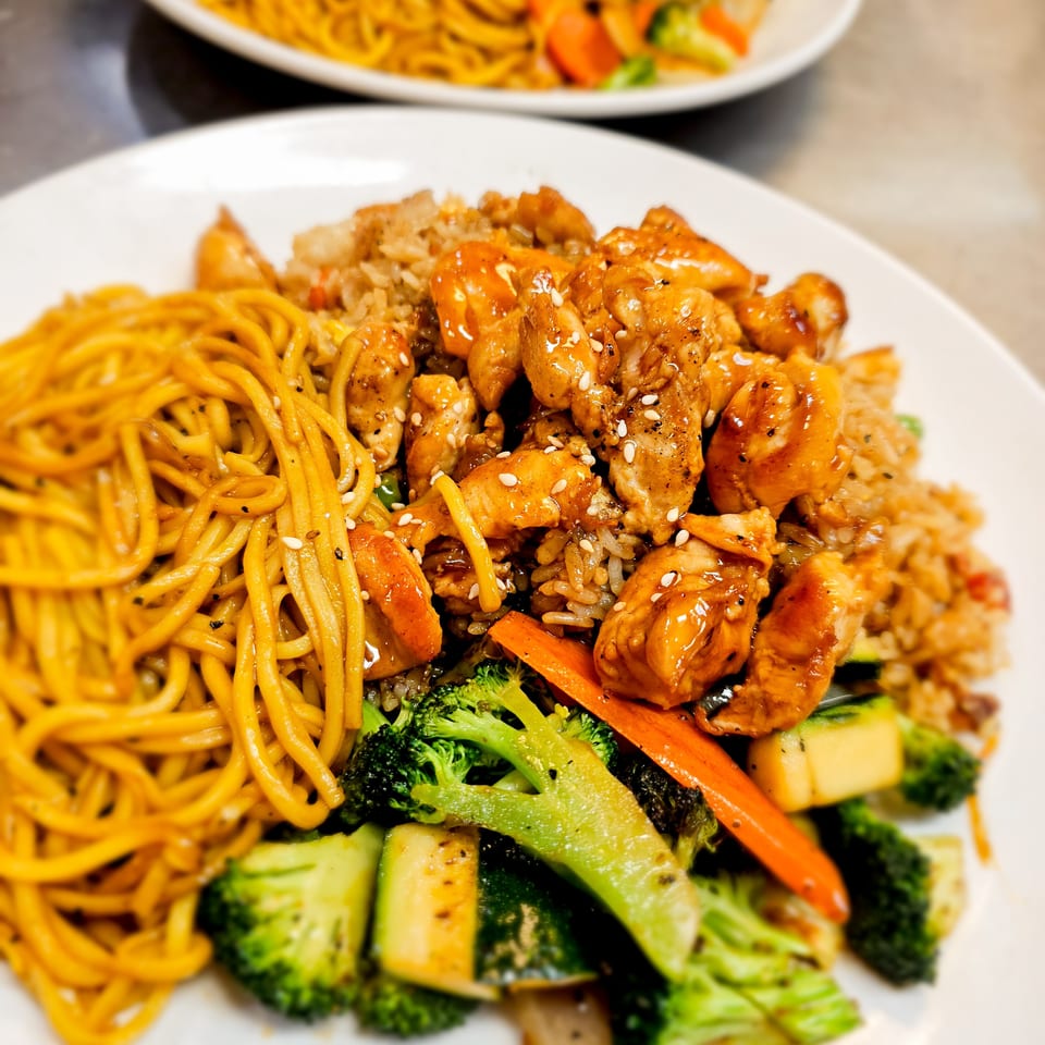 Plate of glazed chicken, fried rice, noodles, and mixed vegetables, with another noodle and grilled meat dish in the background.