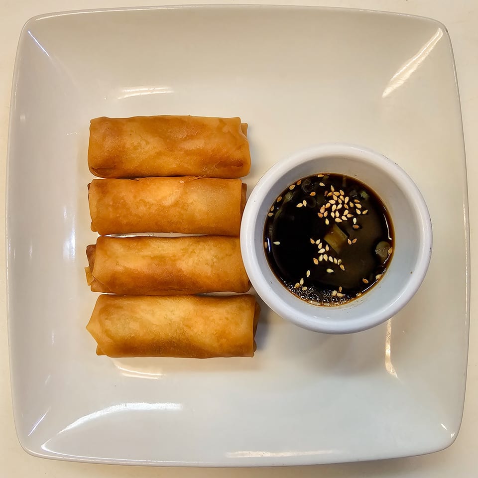 Four golden fried spring rolls served with dark dipping sauce garnished with sesame seeds on a white rectangular plate.