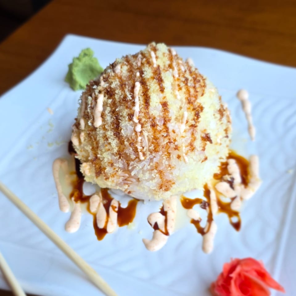 Breaded sushi roll topped with creamy sauce and brown drizzle, served on a white plate with wasabi and a carved tomato garnish.