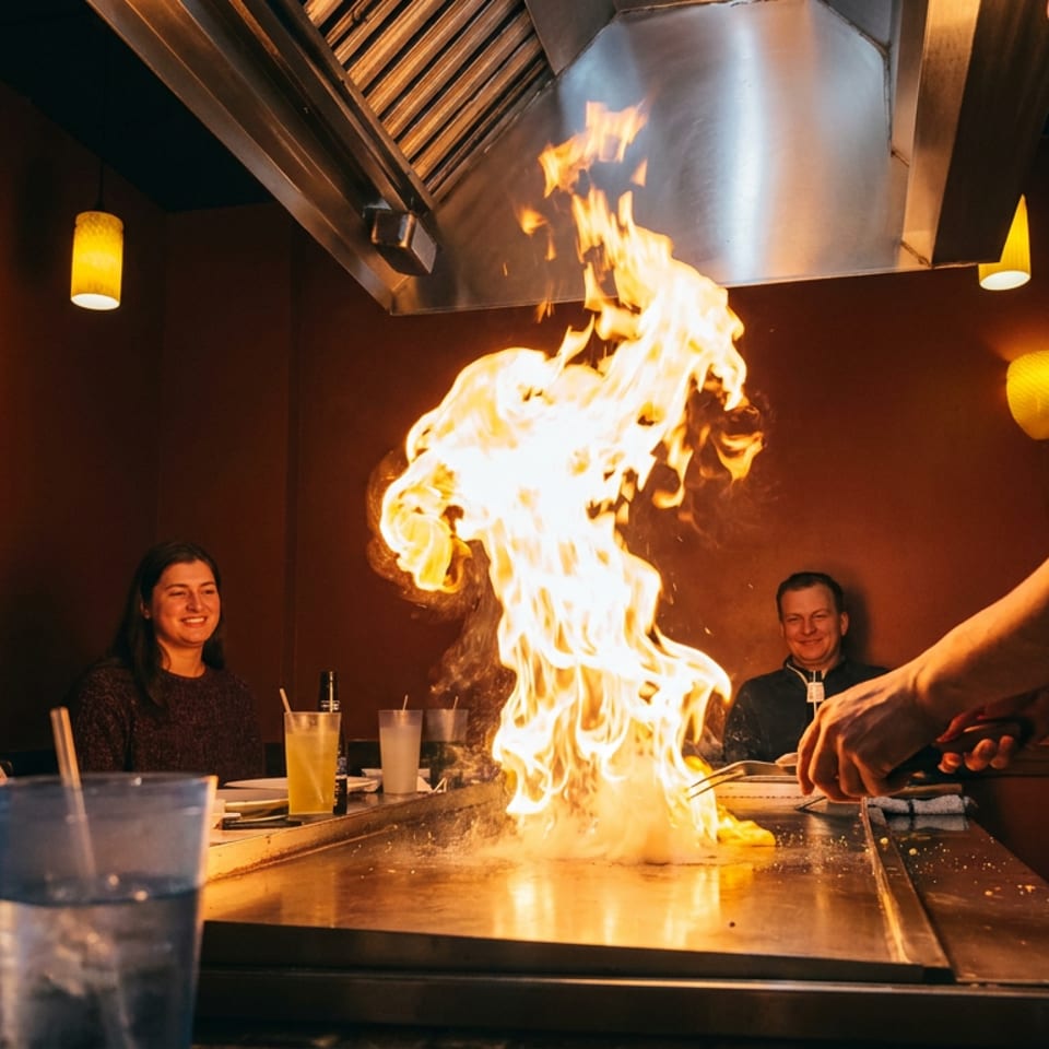 A hibachi chef cooks with a large flame on a grill in front of smiling customers.