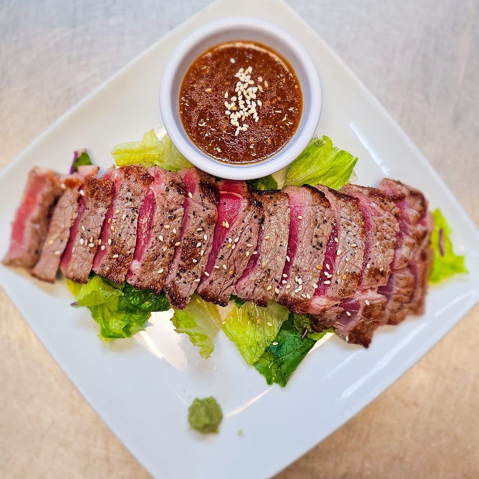 Sliced medium-rare steak garnished with sesame seeds, served over lettuce with spicy dipping sauce on a white plate.